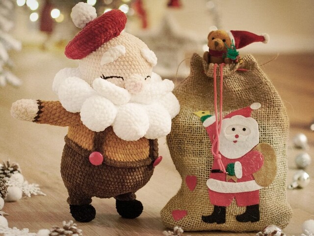 Ein geh&auml;kelter Weihnachtsmann steht in einer Weihnachtslandschaft neben seinem Rucksack mit den Geschenken. Er blickt freundlich und wirkt sehr zufrieden.