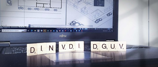vor einem Laptopbildschirm stehen Holzbuchstaben mit den Abkürzungen DIN, VDI und DGUV - zur Artikelseite barrierefreie Wohnungen - Ergänzende Vorschriften
