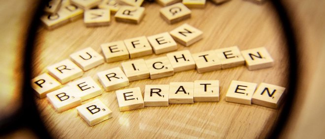 durch eine Lupe sind die Holzbuchstaben mit den Begriffen prüfen, berichten und beraten zu sehen - zur Artikelseite Aufgaben der Überwachungsstelle