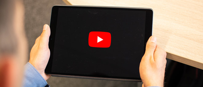 Person hält ein Tablet in der Hand. Es zeigt einen schwarzen Bildschirm mit einem großen roten Button zum Abspielen eines Videos - zur Artikelseite Videos.