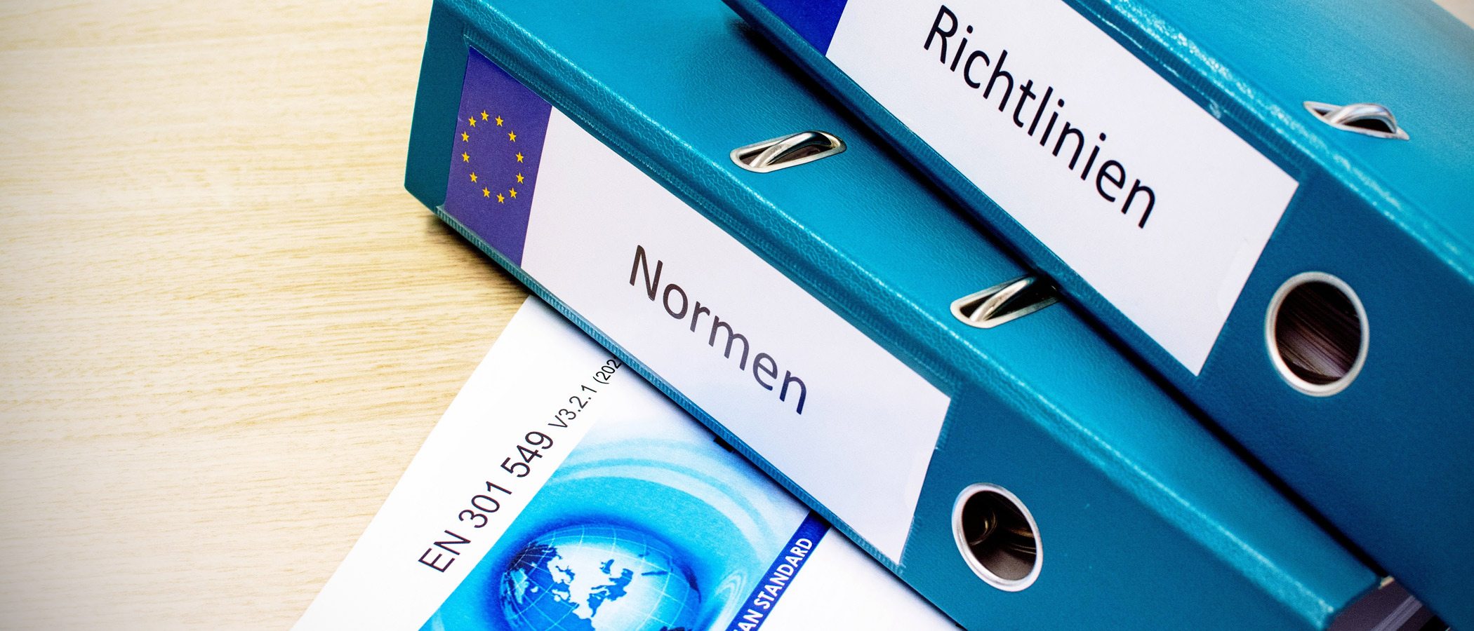 zwei blaue Aktenordner auf einem Schreibtisch mit der Beschriftung Normen und Richtlinien, daneben das Deckblatt der Norm EN 301 549 - zur Artikelseite Normen und Richtlinien
